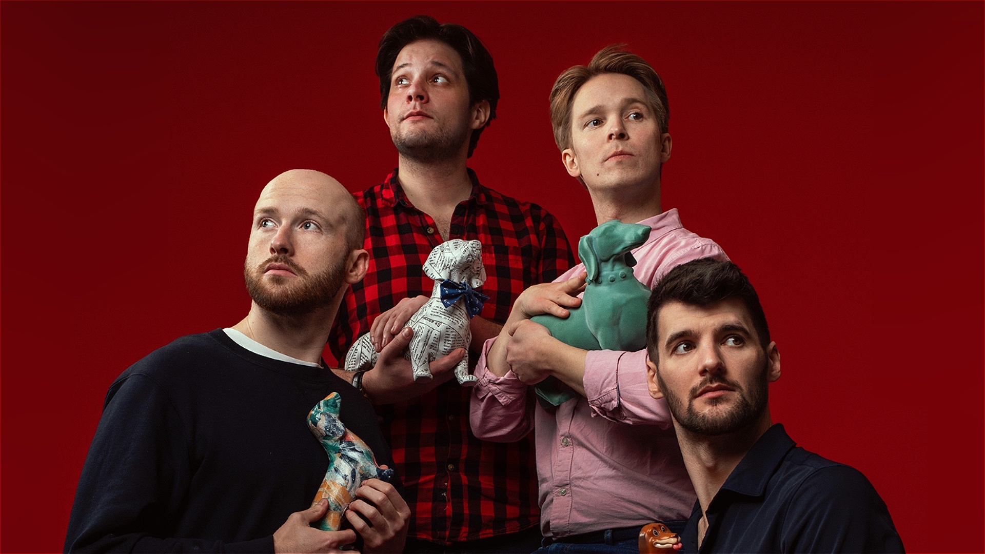 Four adult men pose dramatically against a bright red background. On the left, a bald man with a short reddish beard wears a dark navy sweater and holds a small patterned dog figurine close to his chest. Behind him, a taller man with medium-length dark h