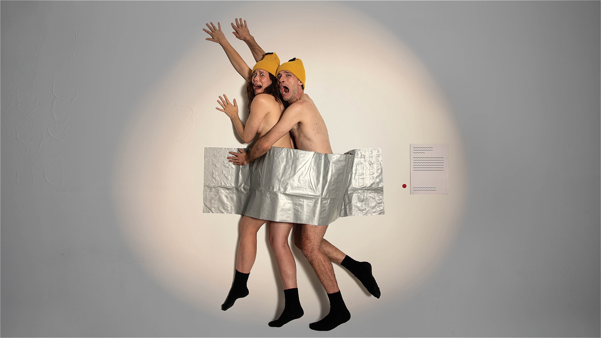 Two actors representing bananas are duct-taped to a white wall, looking afraid.