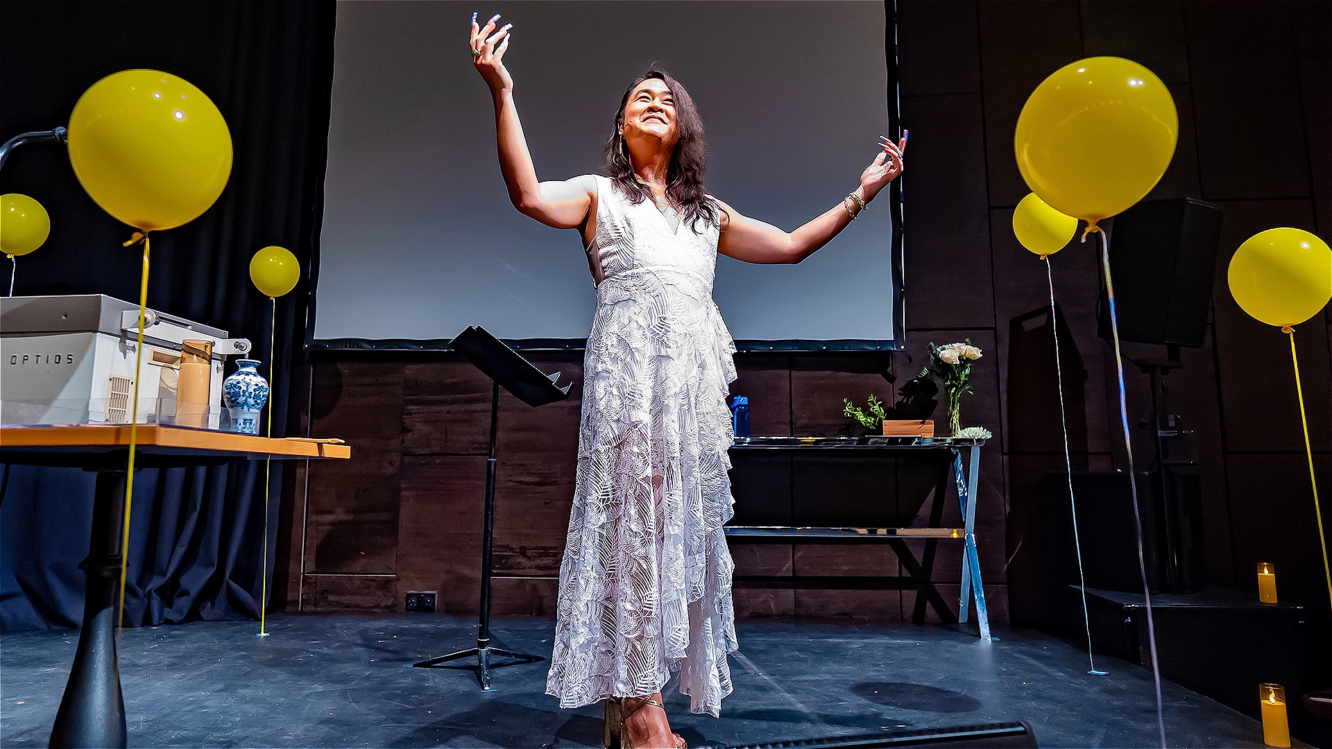 A non-binary person stands in the middle of the stage, gesturing to the crowd, as they are surrounded by yellow floating balloons