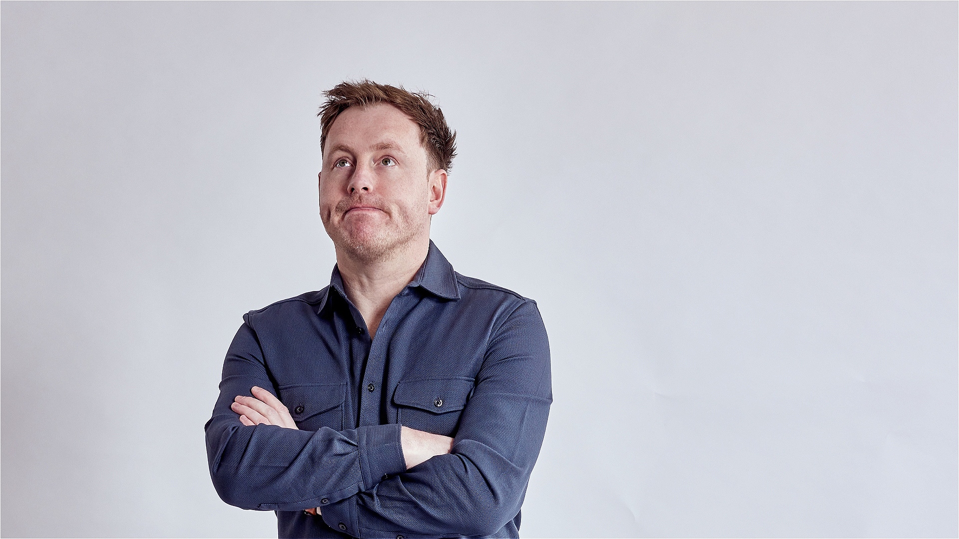 A photo of Andrew Ryan, he crosses his arms and looks upwards with pursed lips. 