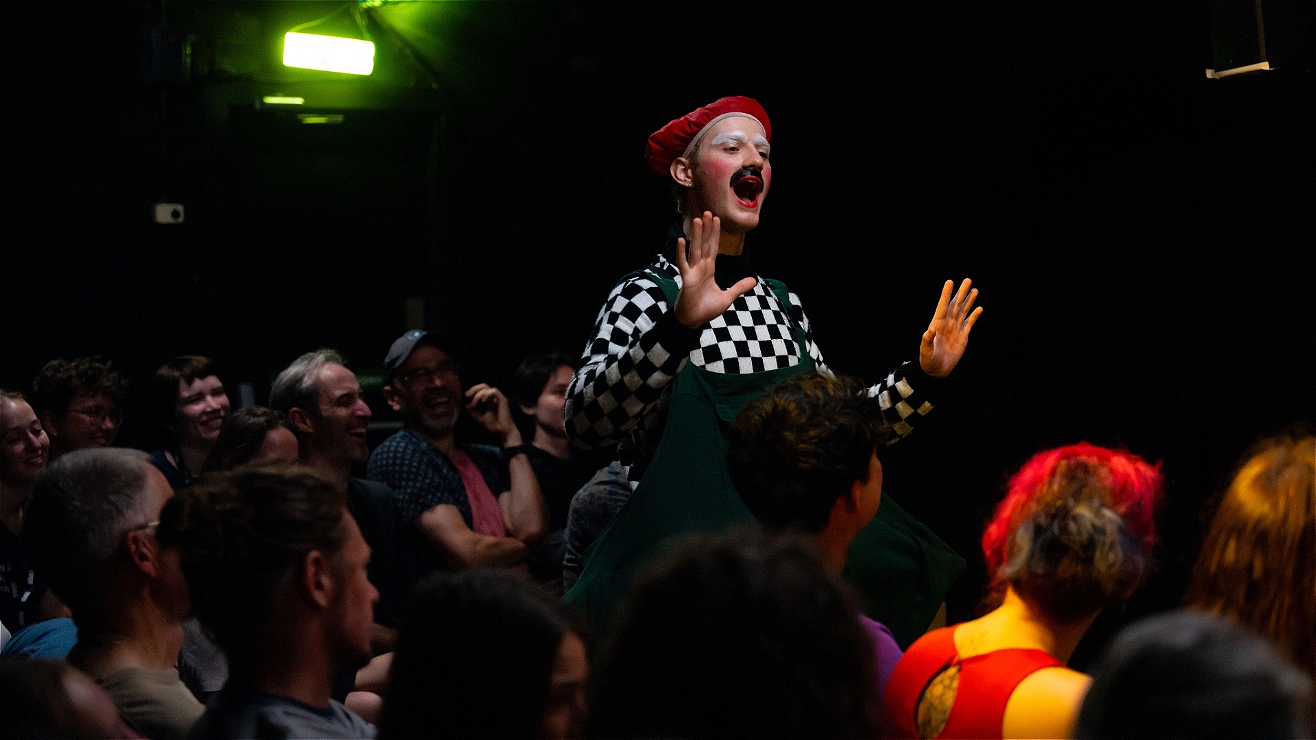 Mr. Cardboard, a colorful imaginary friend in a red cap, wide green overalls, and a checkered sweater, floats above the heads of the seated audiance with his hands indicating for them to calm down as they all are laughing too hard.
