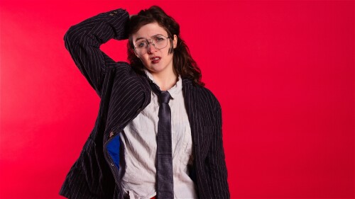 A person dressed in a crumpled white shirt, tie and black jacket stands in front of a red background running one hand through their hair and looking disheveled.