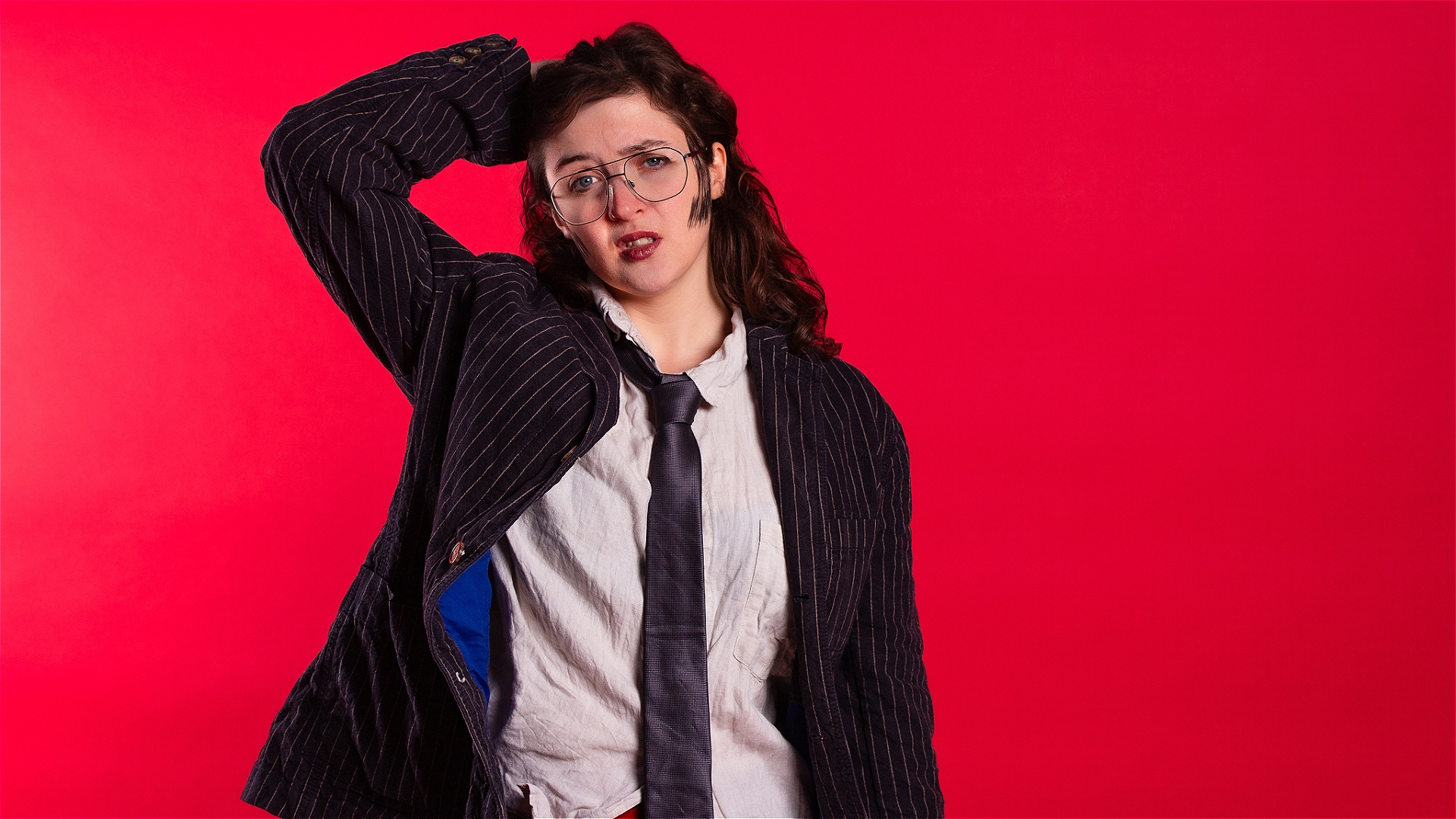 A person dressed in a crumpled white shirt, tie and black jacket stands in front of a red background running one hand through their hair and looking disheveled.