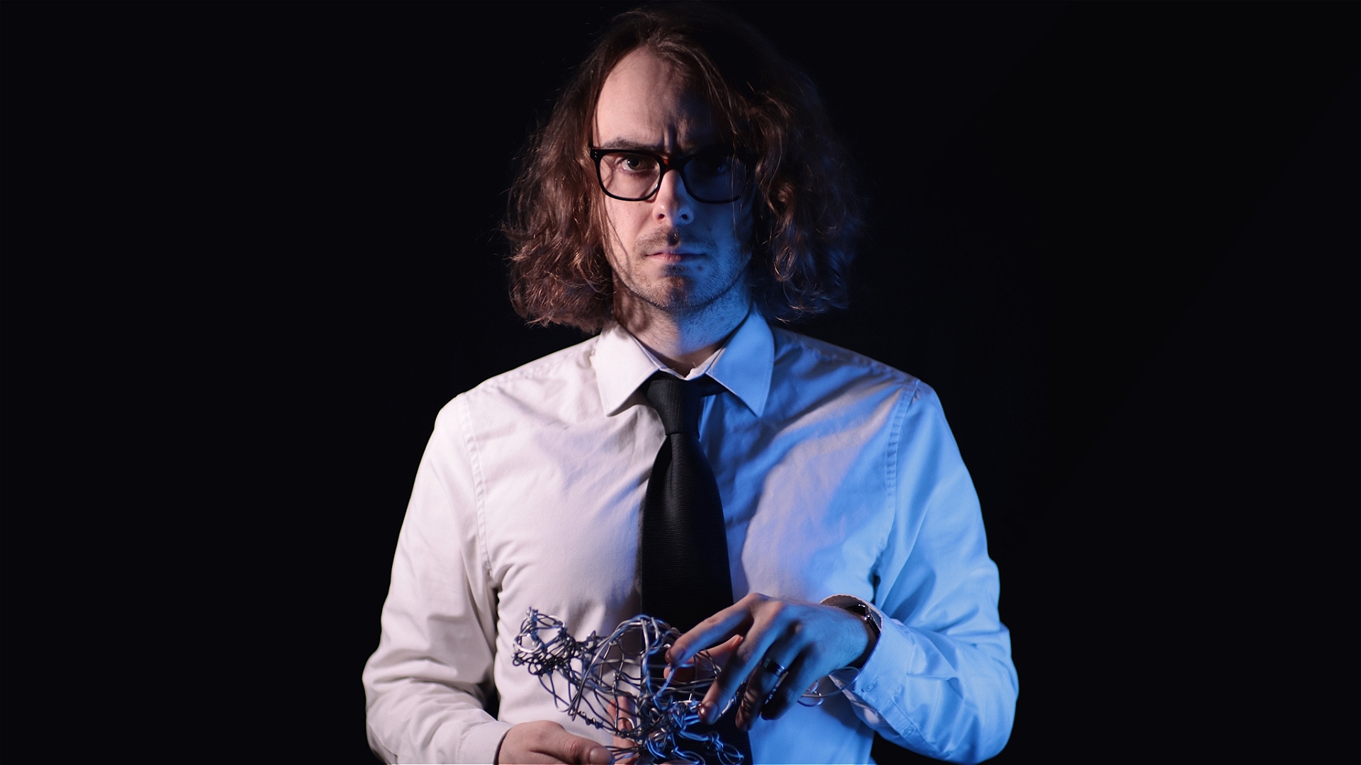A dishevelled, long-haired man wearing glasses, a white shirt, and black tie stares sternly at the camera. He is holding a small wire sculpture of a pigeon in his hands, and is bathed in a blue light, his face partially shadowed, with a black void behind