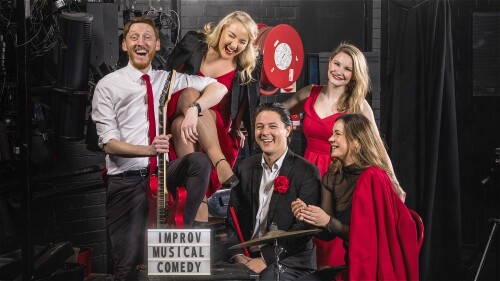 A group of 5 performers holding various musical objects and wearing red and black are laughing and smiling.