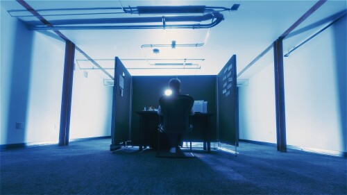 A lone figure sits at a desk in a blue-tinted office cubicle, illuminated by monitor glow, viewed from behind in a cinematic low-angle shot.