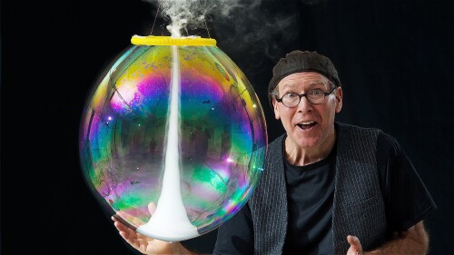 A man in a hat and black clothes holds a large bubble containing a tornado of fog. He looks excited and surprised.