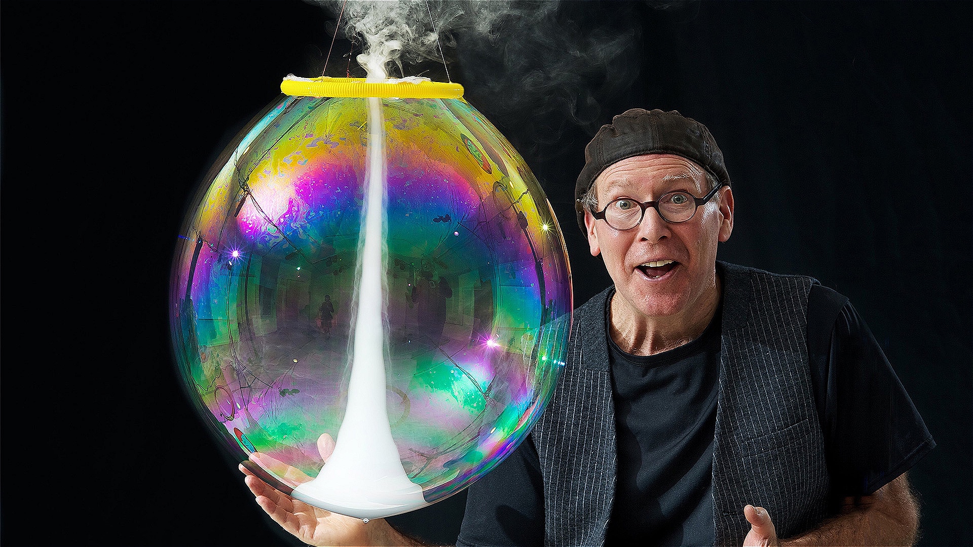 A man in a hat and black clothes holds a large bubble containing a tornado of fog. He looks excited and surprised.