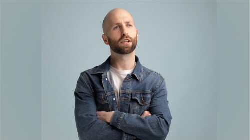 A man in an open denim jacket and white t-shirt sits with his arms crossed