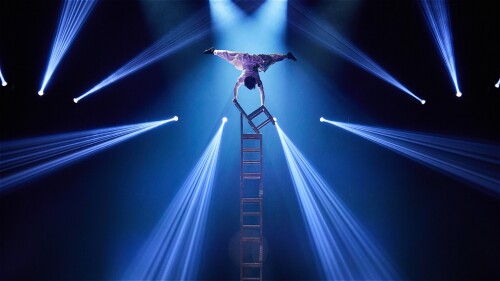 A performer balances on their hands atop a tower of chairs. Streams of light pierce through the darkness to frame them.