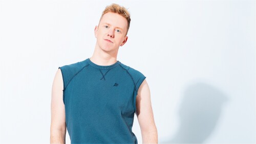 A plain background with a white man posing for the camera. He has blond short hair, and is wearing a turquoise blue tank top and blue jeans.