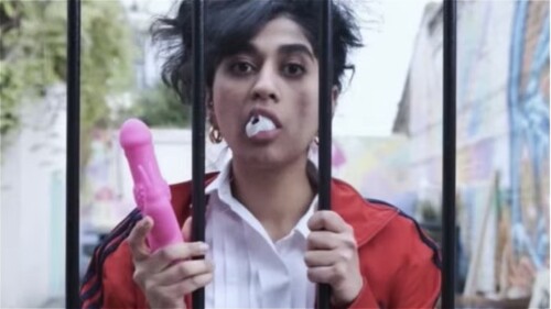 Woman on pink bed, laptop open, she is holding a pink dildo.