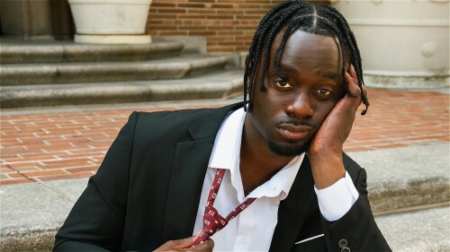 Man wearing a black suit and white dress shirt sits on outdoor stone steps in front of a brick building. He holds a red patterned tie in one hand and rests the side of his face on his other hand.