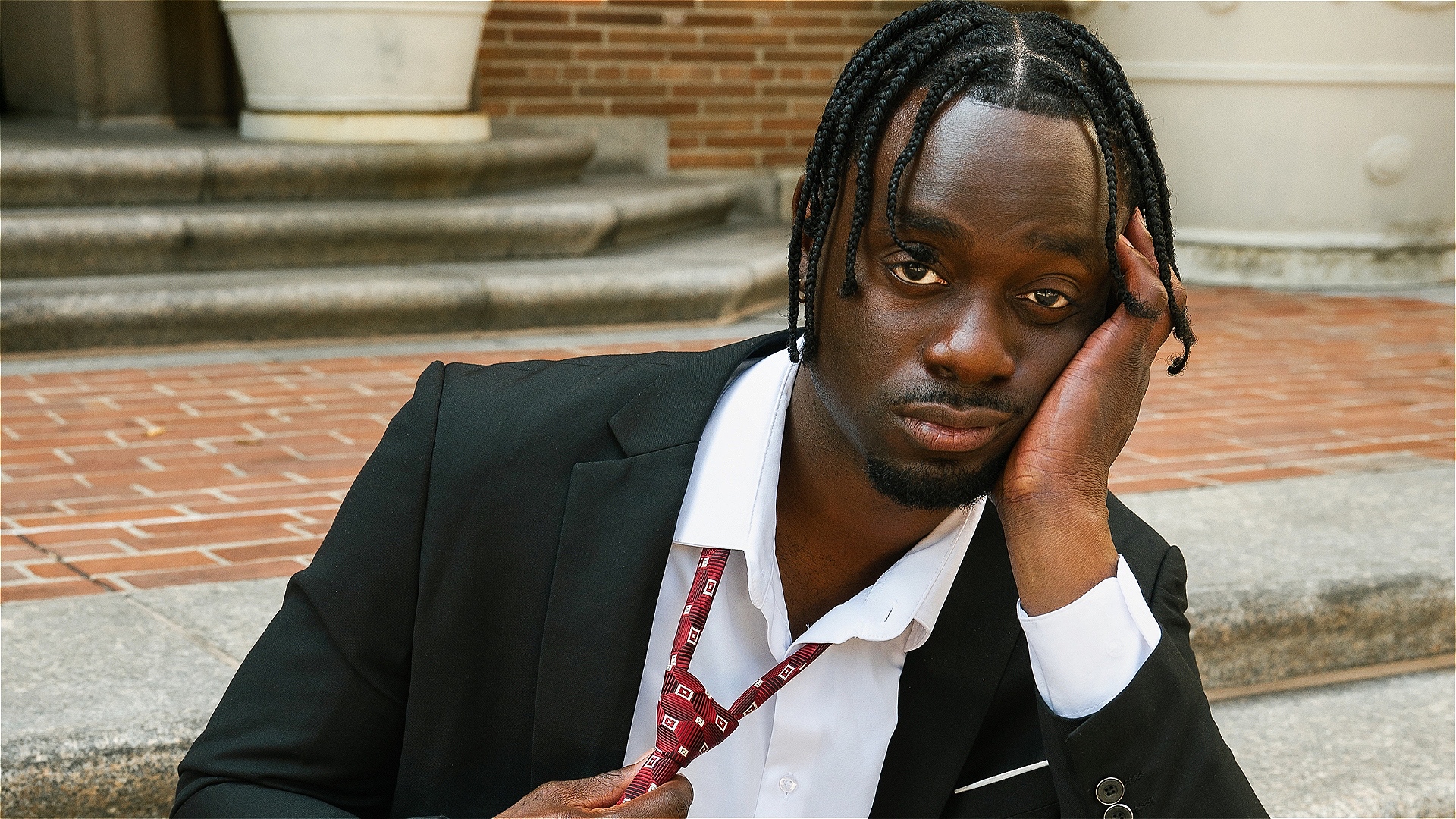 Man wearing a black suit and white dress shirt sits on outdoor stone steps in front of a brick building. He holds a red patterned tie in one hand and rests the side of his face on his other hand.
