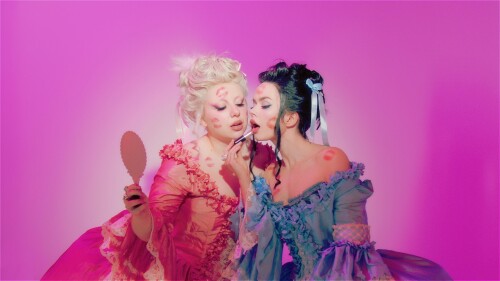 A portrait of Operotica in opulent 18th-century Rococo gowns, covered in lipstick kiss marks. Amber intimately applies lipstick while Dani holds a hand mirror.