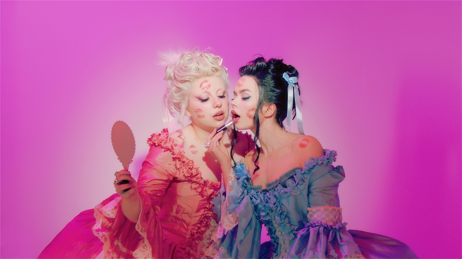 A portrait of Operotica in opulent 18th-century Rococo gowns, covered in lipstick kiss marks. Amber intimately applies lipstick while Dani holds a hand mirror.
