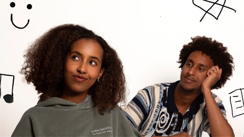 A young boy and girl are sitting at a table facing forward. The girl is looking upward into the air while the boy is looking at her. The girl is wearing a green hoodie, and the boy is wearing a white and blue shirt.