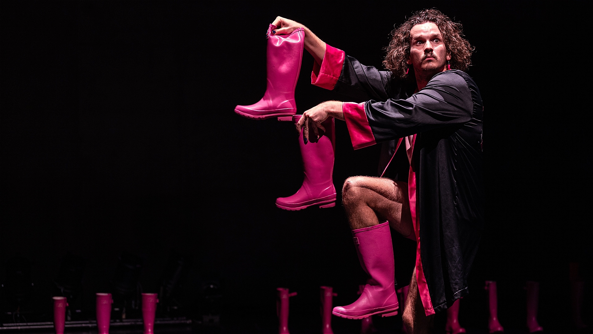 A curly haired performer wearing bright pink boots, a sparkly black and pink gown, and pink boot earrings. He is lifting one foot and holding another pink boot in each hand to make a staircase pattern of boots. He is on a stage full of more pink boots an