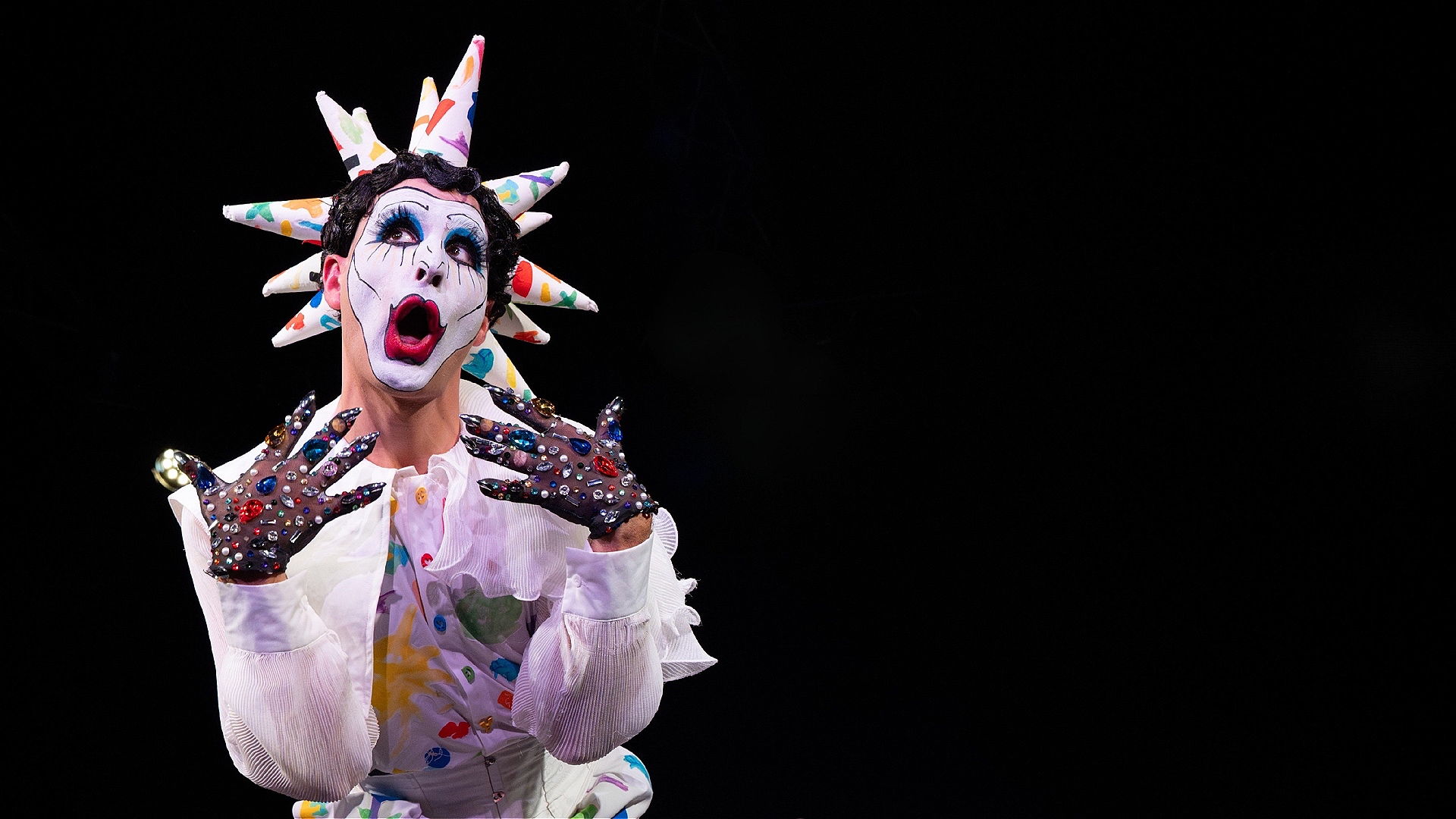 A drag artist in white clown make up and a colourful outfit with a spiked headpiece stands with a shocked look on their face.
