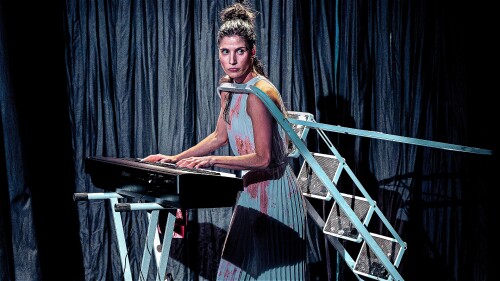 Woman playing a keyboard on stage, wearing a light dress with red stains and a ladder strapped to her back.