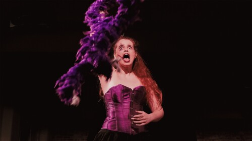 Landscape image: A young woman with glamorous messy clown makeup and long wavy ginger hair is wearing a purple corset and black lace skirt. She is throwing a purple feather boa towards the camera. Her expression is snarling and shocked. Her nails are pai