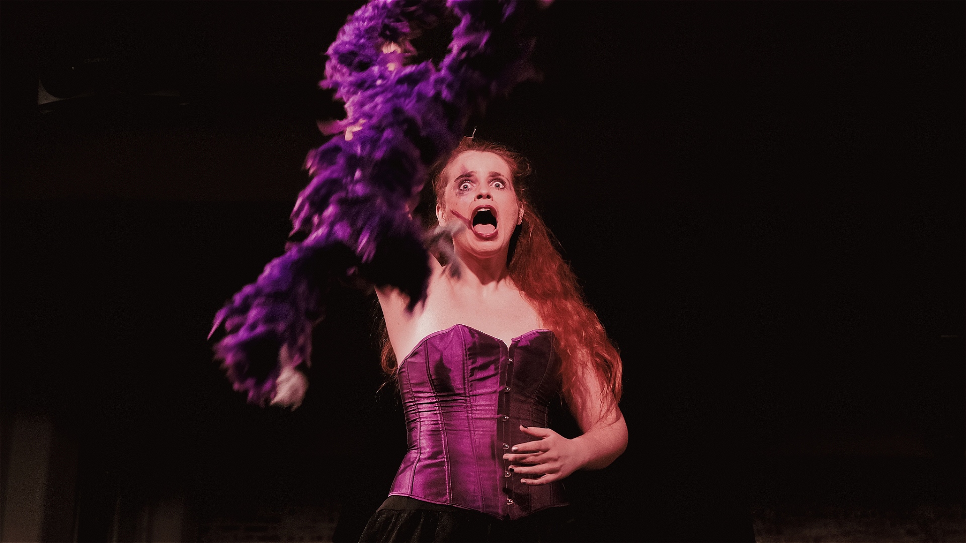 Landscape image: A young woman with glamorous messy clown makeup and long wavy ginger hair is wearing a purple corset and black lace skirt. She is throwing a purple feather boa towards the camera. Her expression is snarling and shocked. Her nails are pai