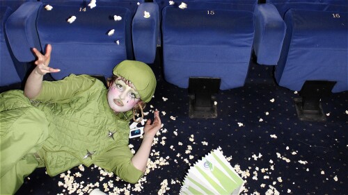 A person in an all-green outfit and beret lies on the floor in a cinema in a pile of spilled popcorn