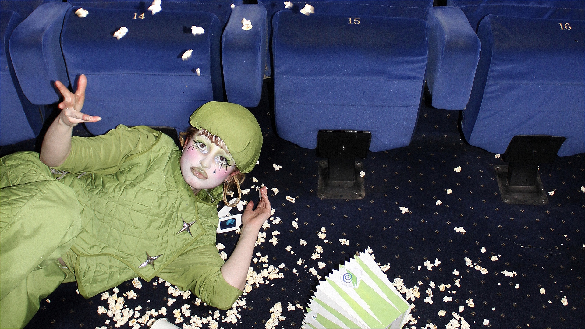 A person in an all-green outfit and beret lies on the floor in a cinema in a pile of spilled popcorn