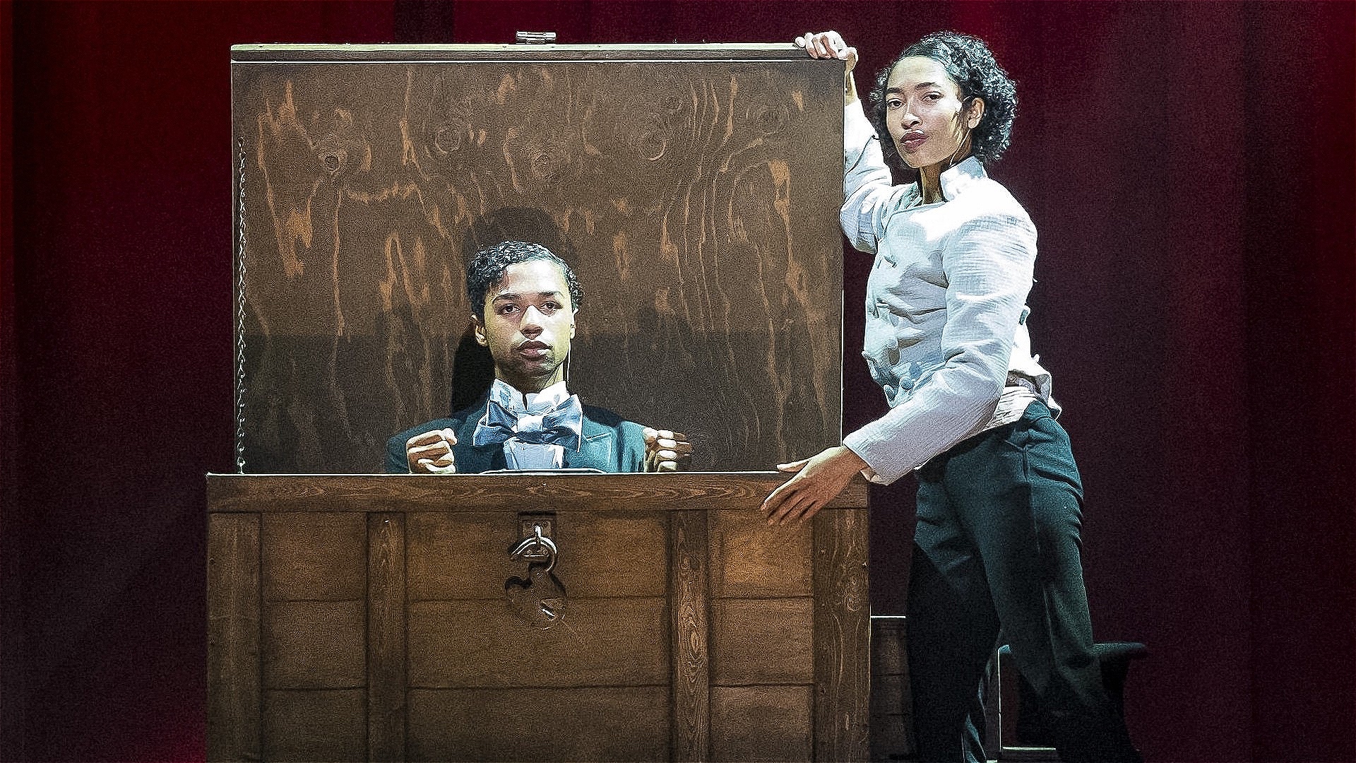 Man in a tailcoat kneeling in a woden chest with a woman about to close the lid