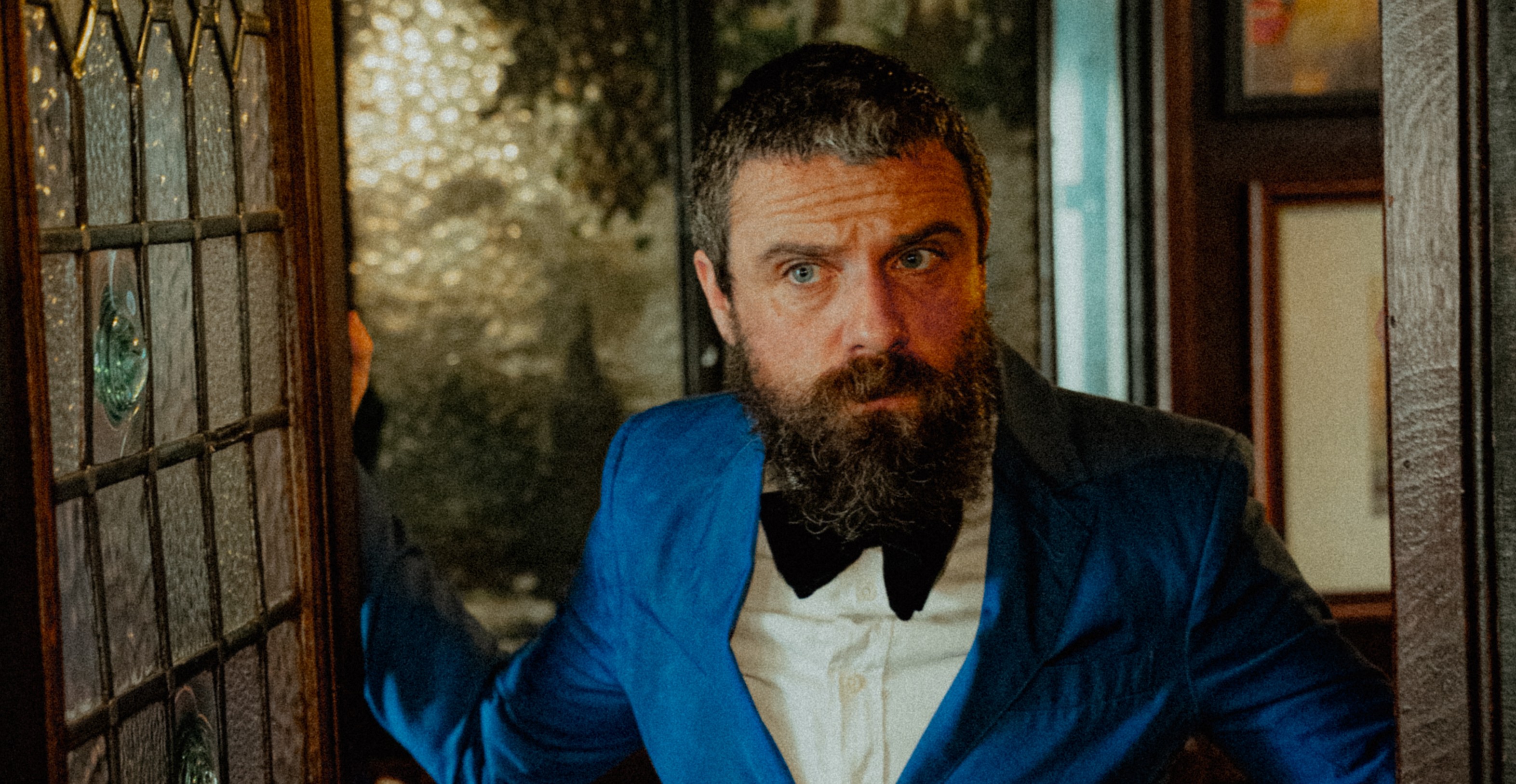a man in a blue suit, white shirt and black bow tie stands opening a front door