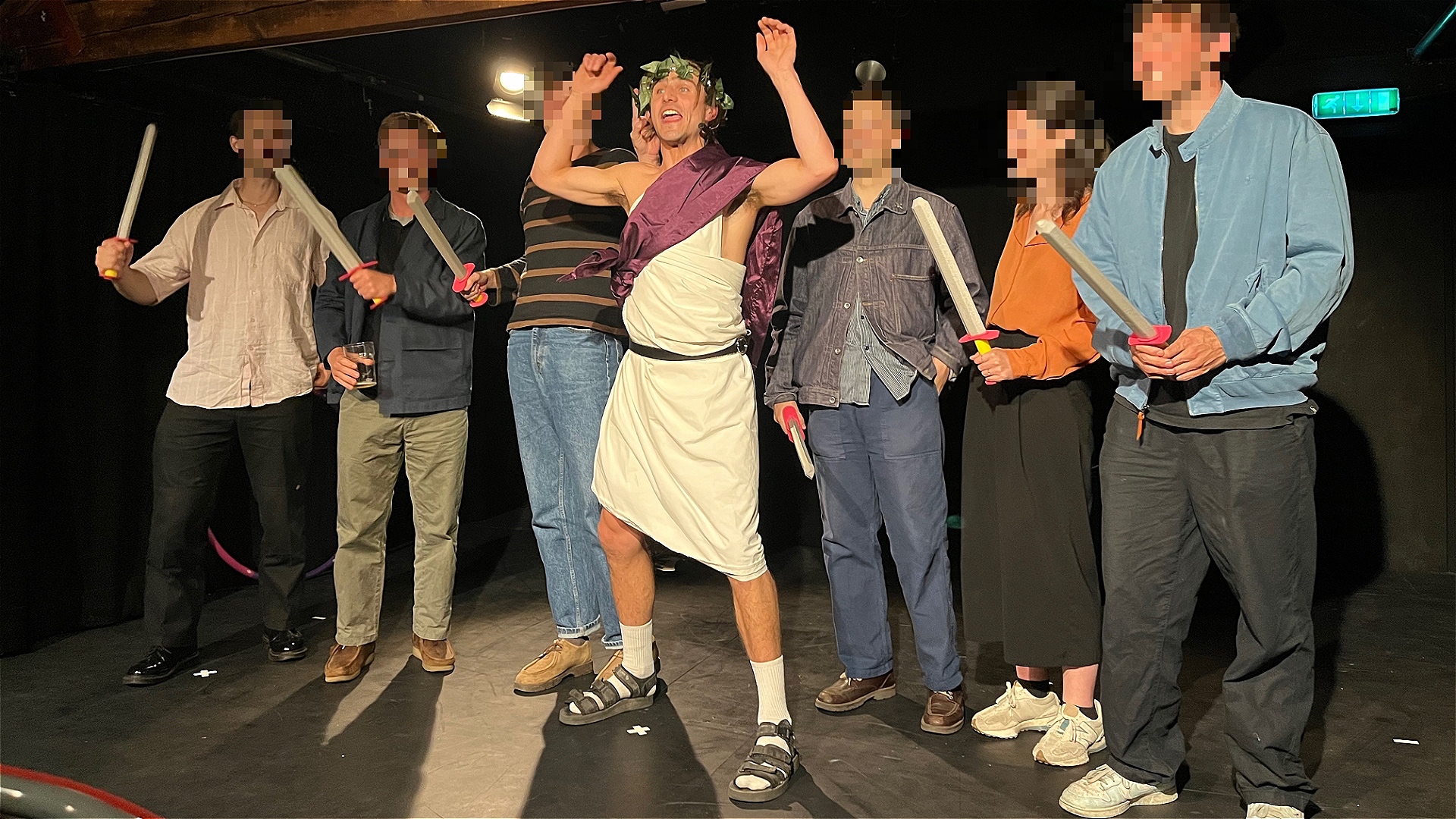 A techbro Julius Caesar stands with arms raised in the middle of a group of six people with faces blurred holding foam daggers.