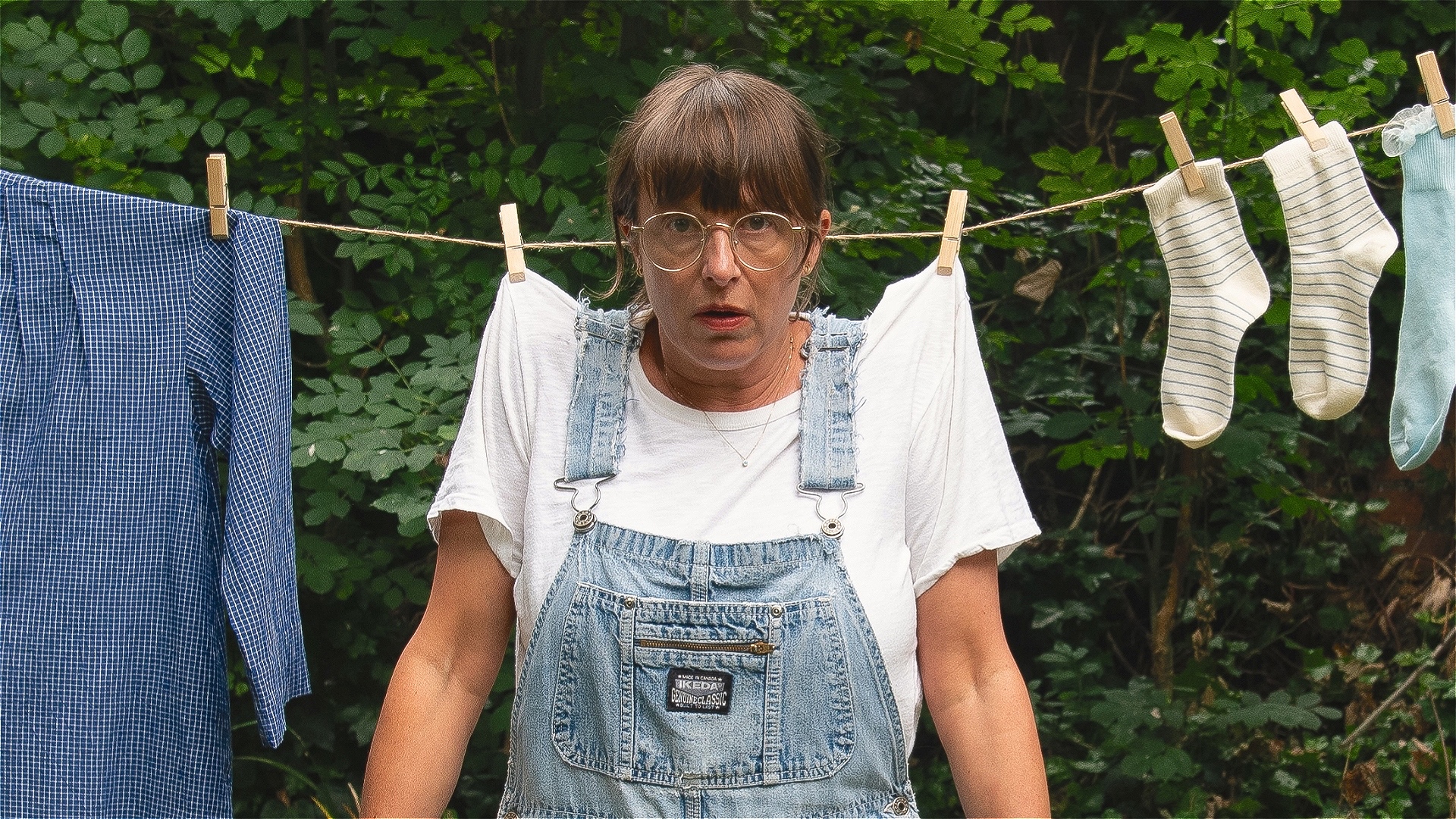 Bonnie Oddie, writer and performer, wears dungarees and is hung from a clothes line by two clothes pegs. There are socks and a shirt on the line next to her