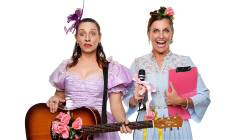 1 woman with brown hair and a worried look on her face, wearing a lavender bridesmaid dress, holding an acoustic guitar with a bunch of flowers coming out of the sound hole, and a 2nd woman with an over excited look on her face, brown hair, holding a pin