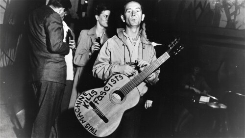 Woody Guthrie backstage with a cigarette and his guitar which has the words 