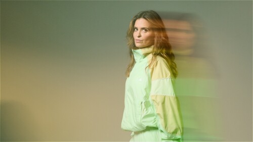 A woman in a light green tracksuit stands holding a retro boombox, with motion blur creating a ghosted double image beside her against a soft, muted background.