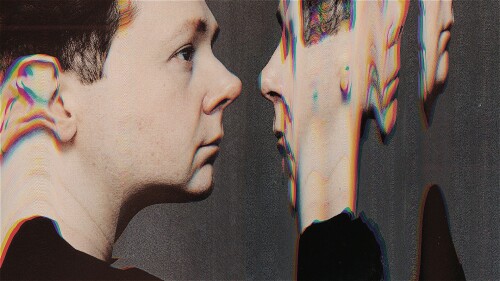 Profile view of a man facing his own digitally distorted reflection, with chromatic aberration and glitch effects warping the mirrored face against a dark background.
