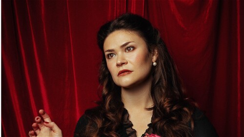 Photograph of woman standing in front of red velvet curtain. She is holding a bouquet of pink and white flowers in one hand and the other hand is raised as if about to make a gesture. She is gazing into the distance.