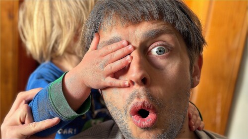 A man looks shocked while a toddler's small hand covers his right eye