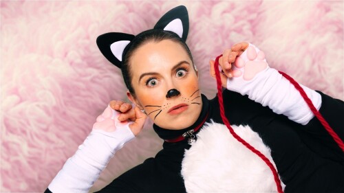 A woman dressed as a cat, with black and white ears, white gloves with pink paws on the palm, black turtle neck with white fur patch on the chest and a red collar around the neck, lays on her side with paws beside her face and a scared/surprised expressi