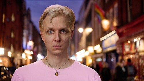 The face of Linus Karp (a blonde man with blue eyes) is looking directly at you. He's wearing a pink t-shirt and a necklace with an image of Princess Diana. He's standing against a background of a blurred street in Soho, London.