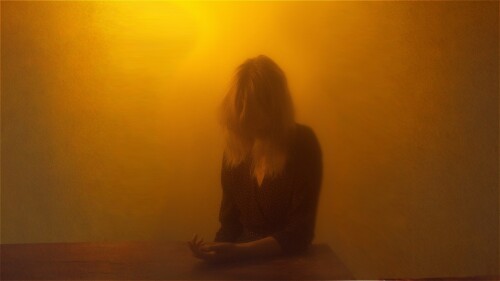 A softly blurred, warm-toned image shows a person sitting at a table with their head slightly bowed. Their medium-length hair falls forward, partially covering their face. The lighting is dim and orange-hued, creating a moody, atmospheric effect, and the