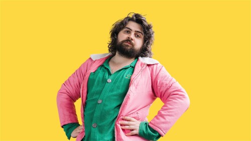 Yoz Mensch is looking directly at the camera, standing with both hands on her waist in front of a bright yellow seamless background. Yoz is a white person, with 70's style shaggy shoulder length dark brown hair, thick eyebrows and a thick beard. The expr