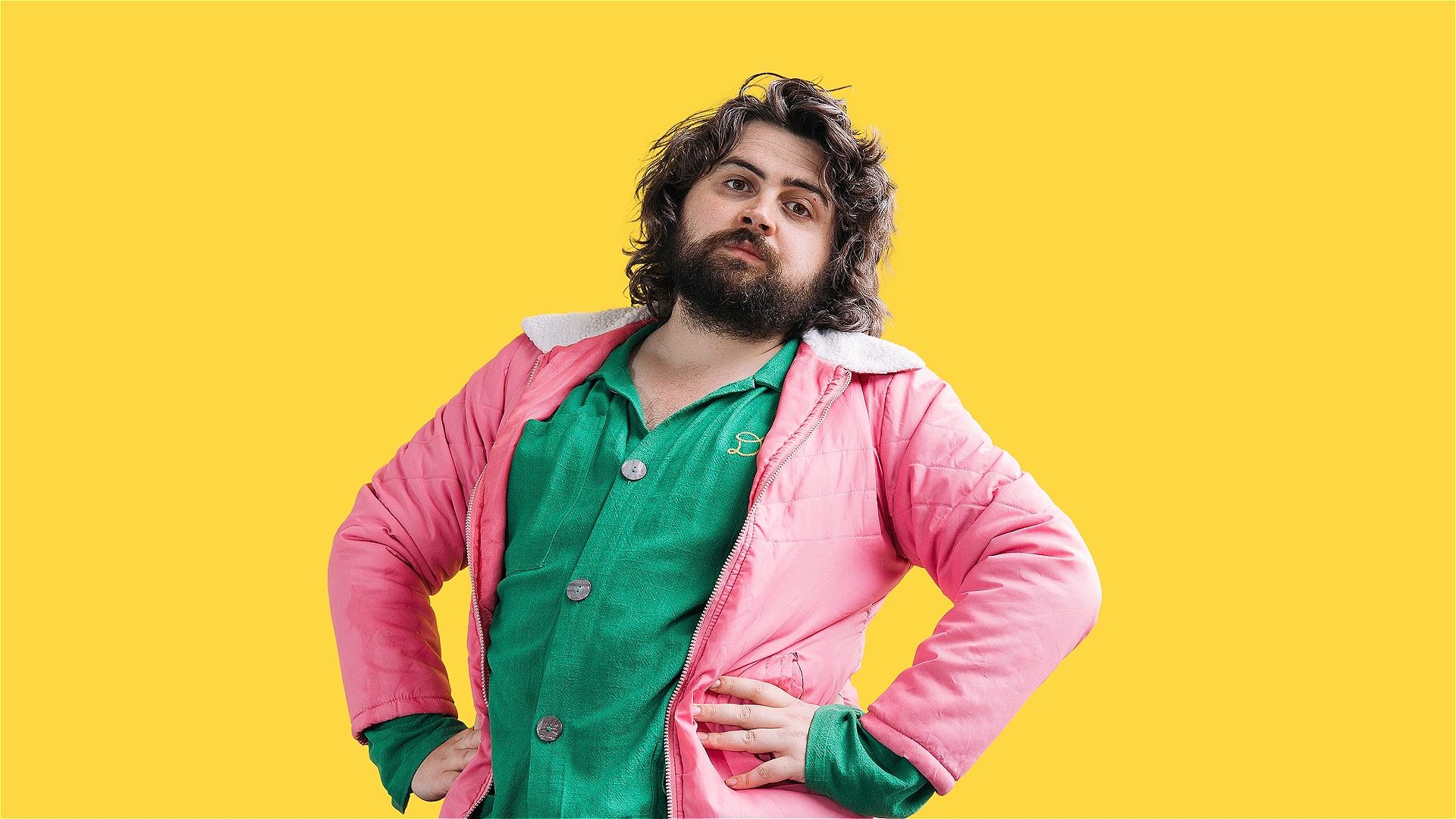 Yoz Mensch is looking directly at the camera, standing with both hands on her waist in front of a bright yellow seamless background. Yoz is a white person, with 70's style shaggy shoulder length dark brown hair, thick eyebrows and a thick beard. The expr