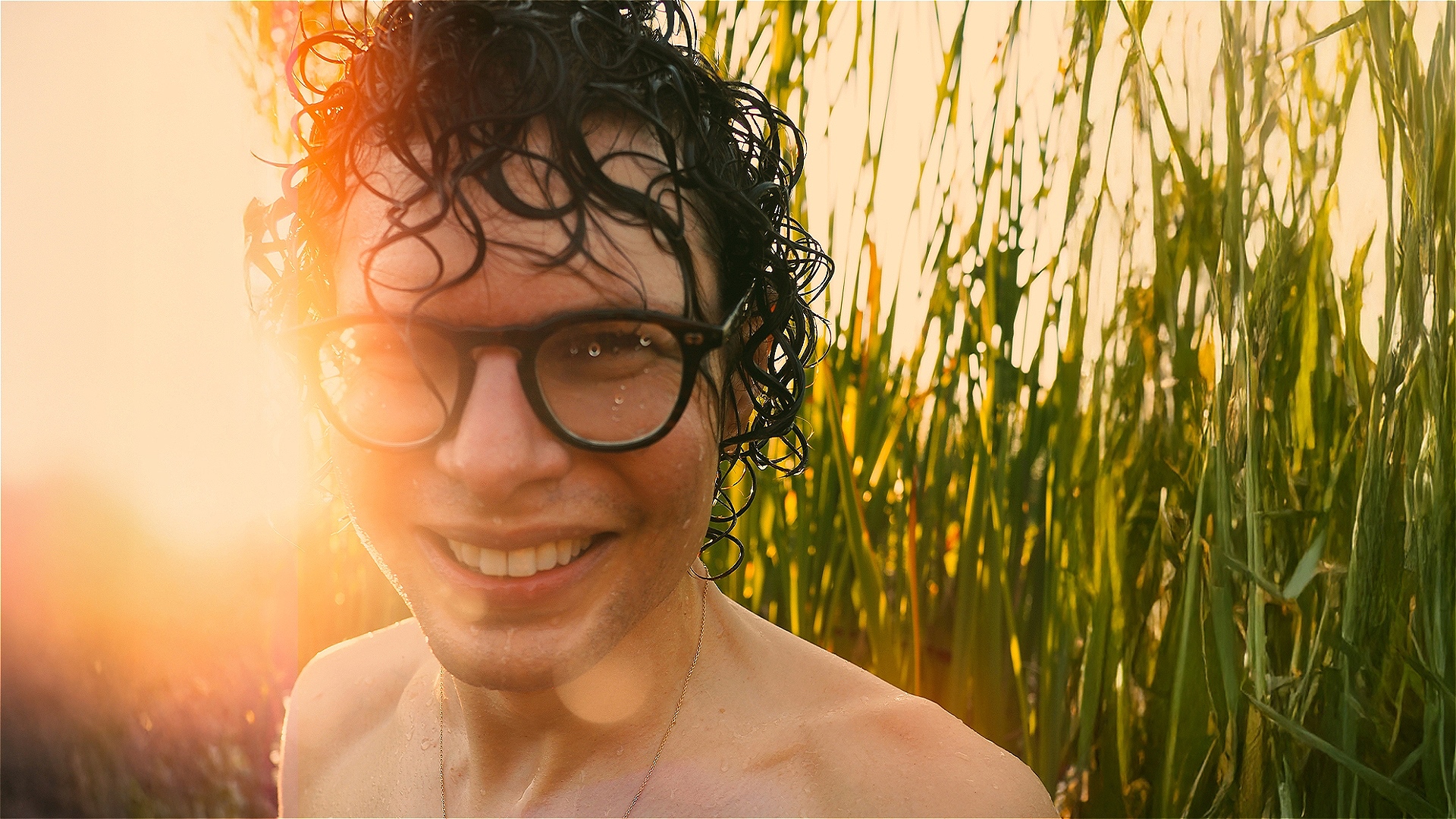 A man sits topless in a field with the sun shining on him, his hair is tousled and he smiles to camera