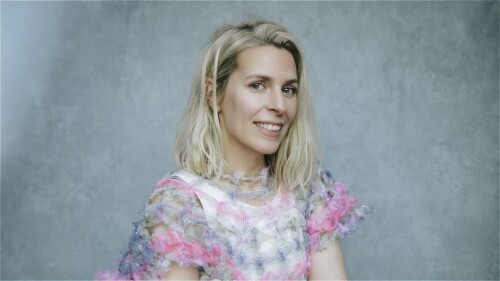 Sara, a blonde white woman wearing a black and white top, smiles at the camera against a grey background.