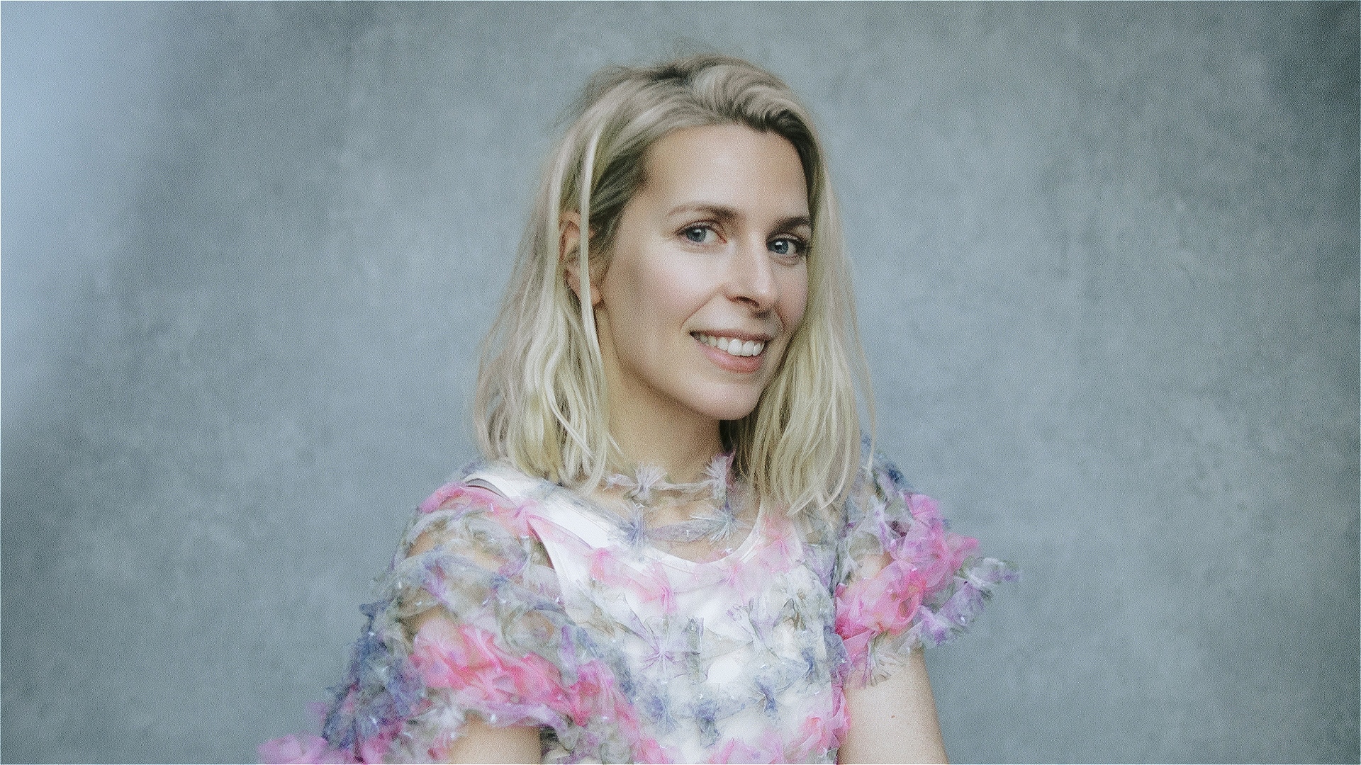 Sara, a blonde white woman wearing a black and white top, smiles at the camera against a grey background.