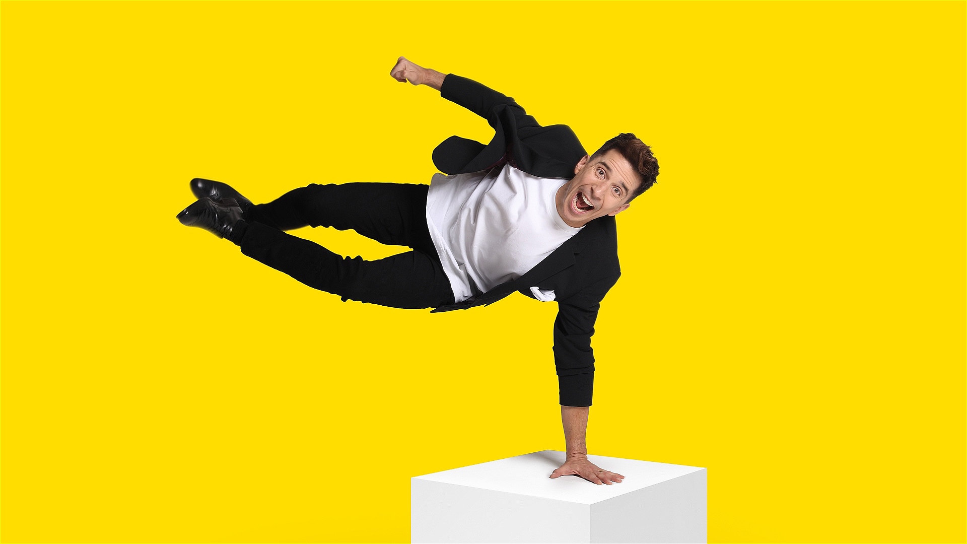 A man in a dark suit and white t-shirt jumps into the air with his hand on a white block against a yellow background