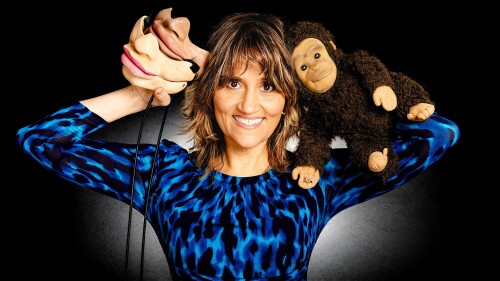 A female performer stands in a dazzling blue dress holding a monkey puppet in one arm and a pair of ventriloquist's face masks in the other arm.