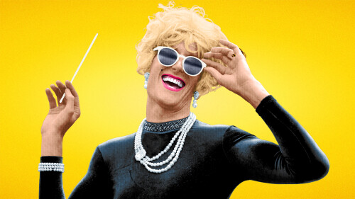 A blonde drag queen with white sunglasses in a black dress poses against a yellow background