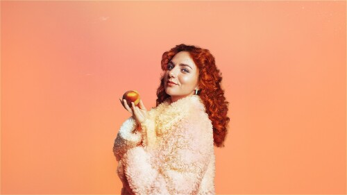 Comedian Ania Magliano holding a peach in a fluffy coat, against an orange background